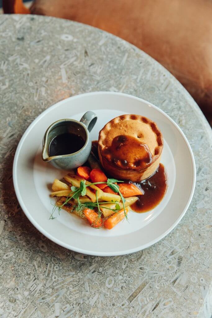 Pie, gravy and vegetables on a white plate, placed on a grey table top