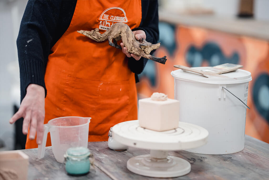 Woman in pottery studio with pottery object, jug with coloured slip to decorate it and white bucket with clay in. Holding a fox sculpture.