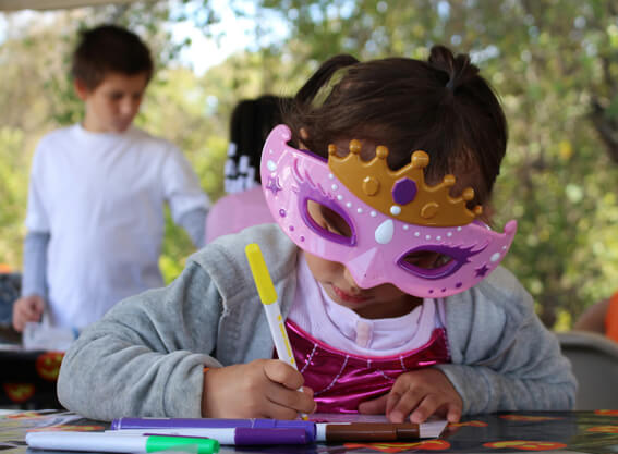 Little girl wearing a pink and gold mask, doing colouring at a table